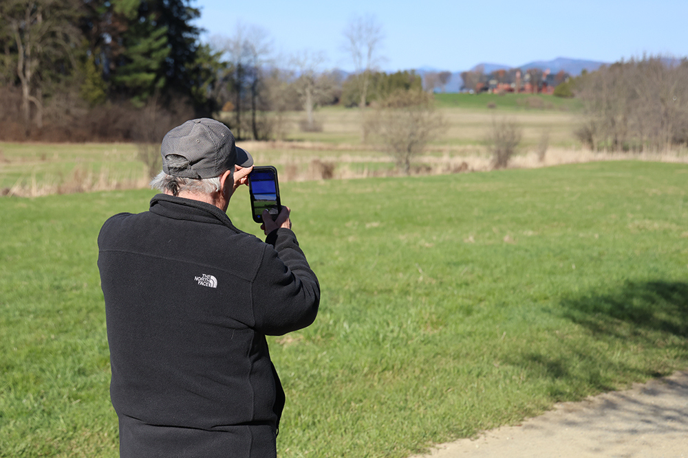 man in baseball cap and black fleece jacket facing a field holding up his camera taking a photo