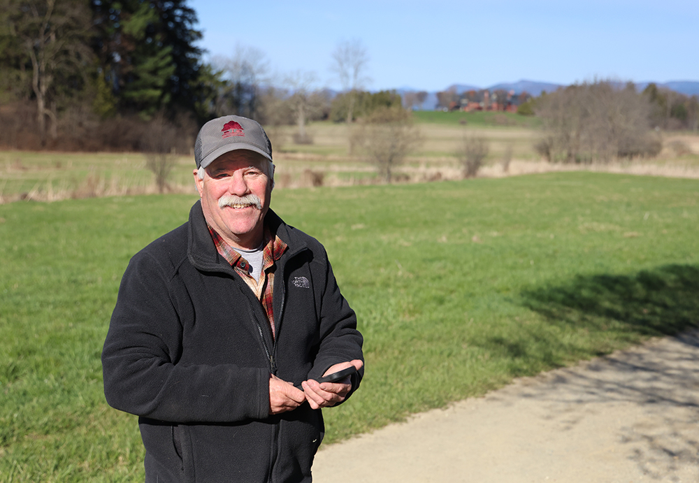 man with mustache and baseball cap in a black fleece jacket smiles at camera. he's holding his cell phone, and there are green fields and sky behind him.