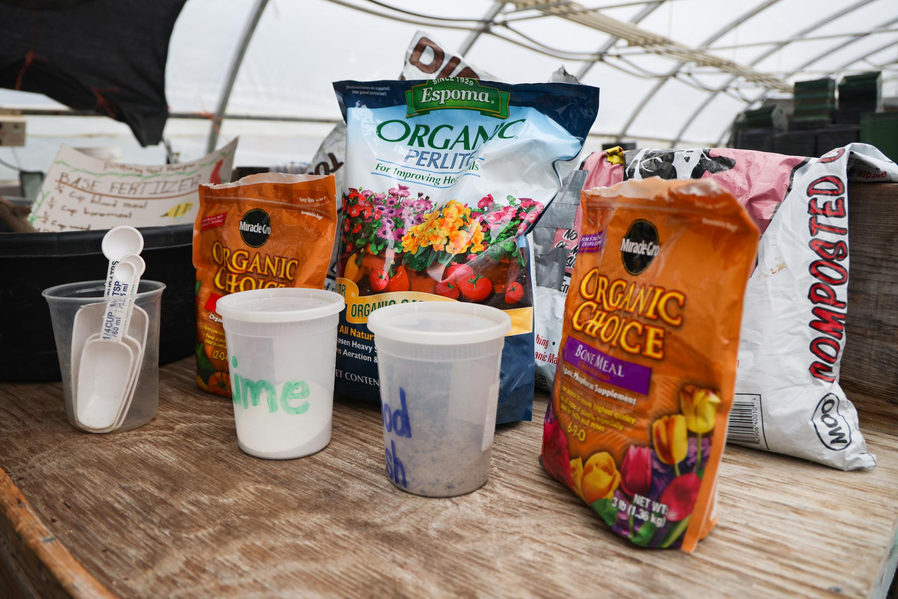Bags of potting mix are on a wooden table in a greenhouse. Small containers labeled with different types of soil and scoopers are also visible.