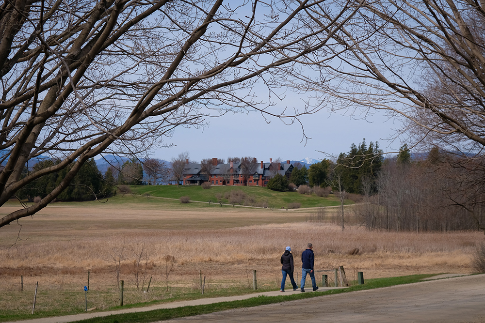 two people walking on trail with tree branches in foreground and inn in background
