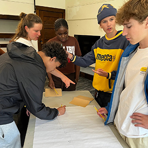 Five young people gather around a table with a large sheet of paper