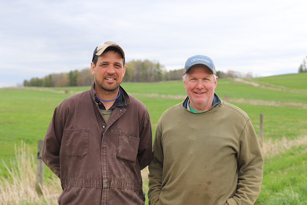 two men standing side by side. man on left is wearing brown coveralls and billed cap, man on right in ragged sweatshirt, gray hair, and blled cap