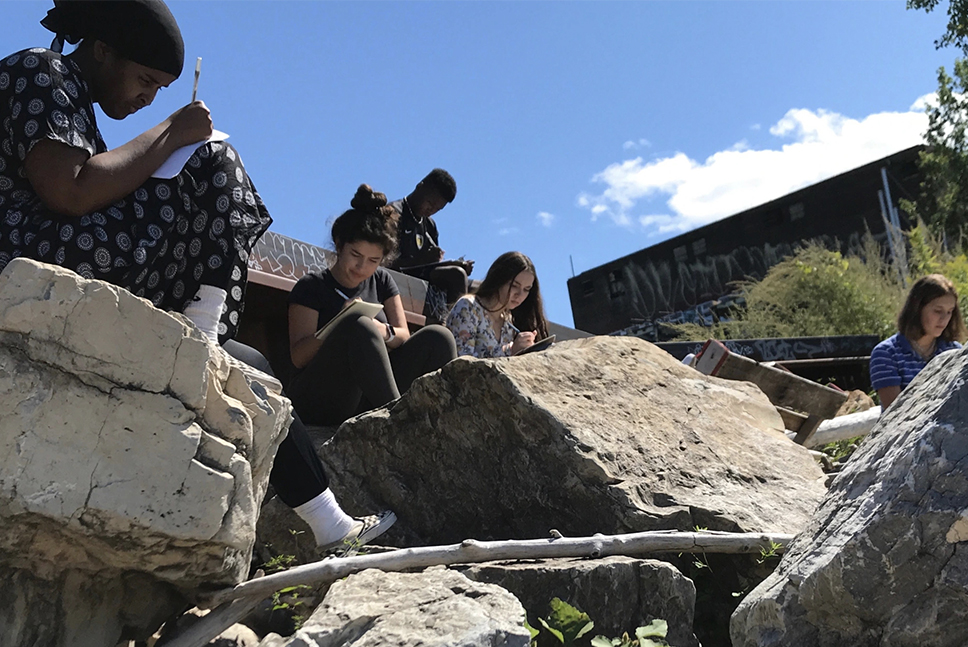Five high school aged students write in journals gathered on a rocky shoreline outside in summertime