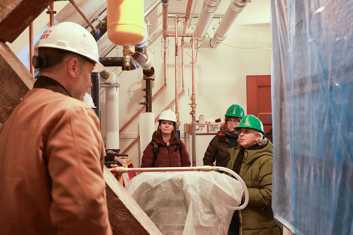 three women in hard hats listening to a man in a hard hat, all in a small room with lots of exposed pipes