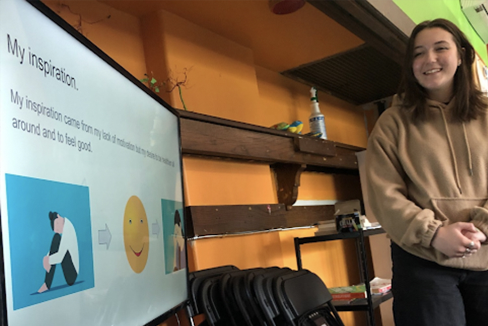 A high school student smiles standing next to a screen displaying a presentation titled My Inspiration