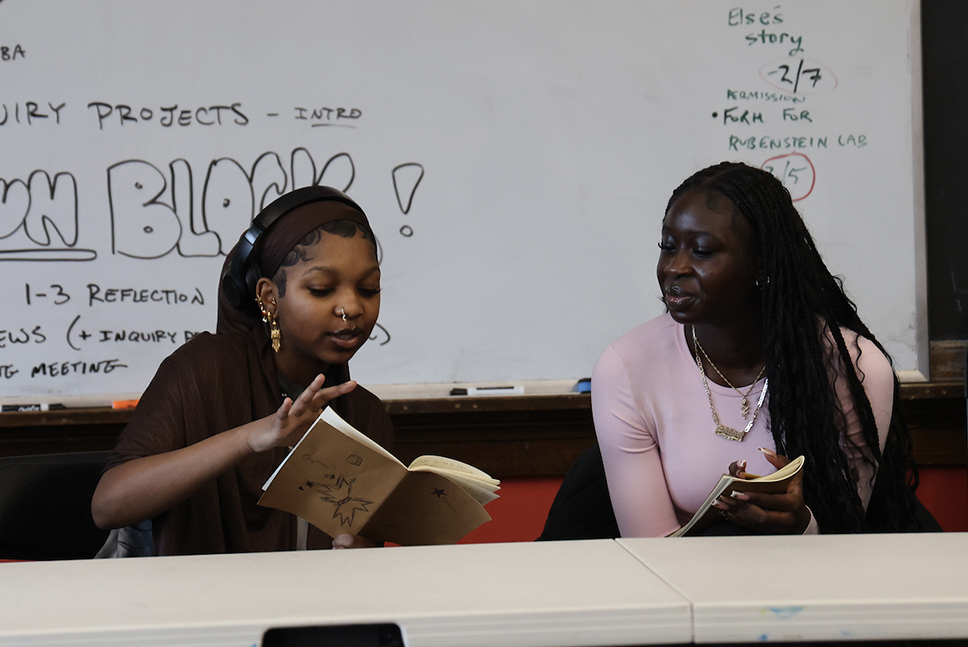 Two high school students share journals while sitting in a classroom in front of a whiteboard