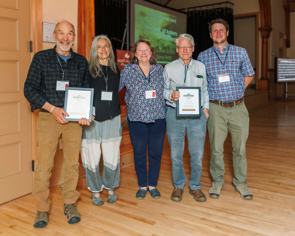 5 people standing in a group smiling at the camera. The first and fourth people are each holding an award plaque.
