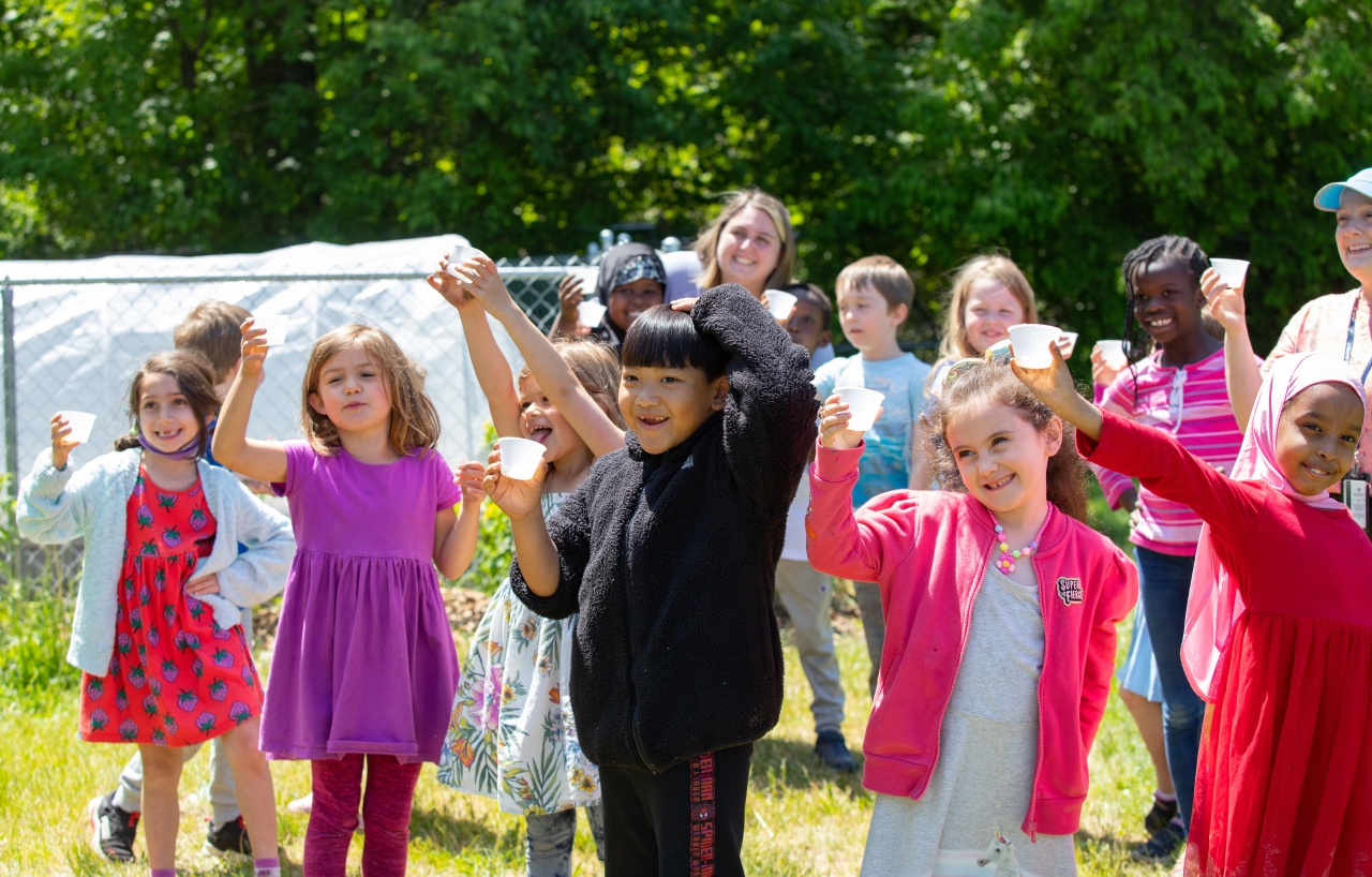 A group of a dozen children and adults smile while raising small paper cups on a sunny outdoor grassy lawn.