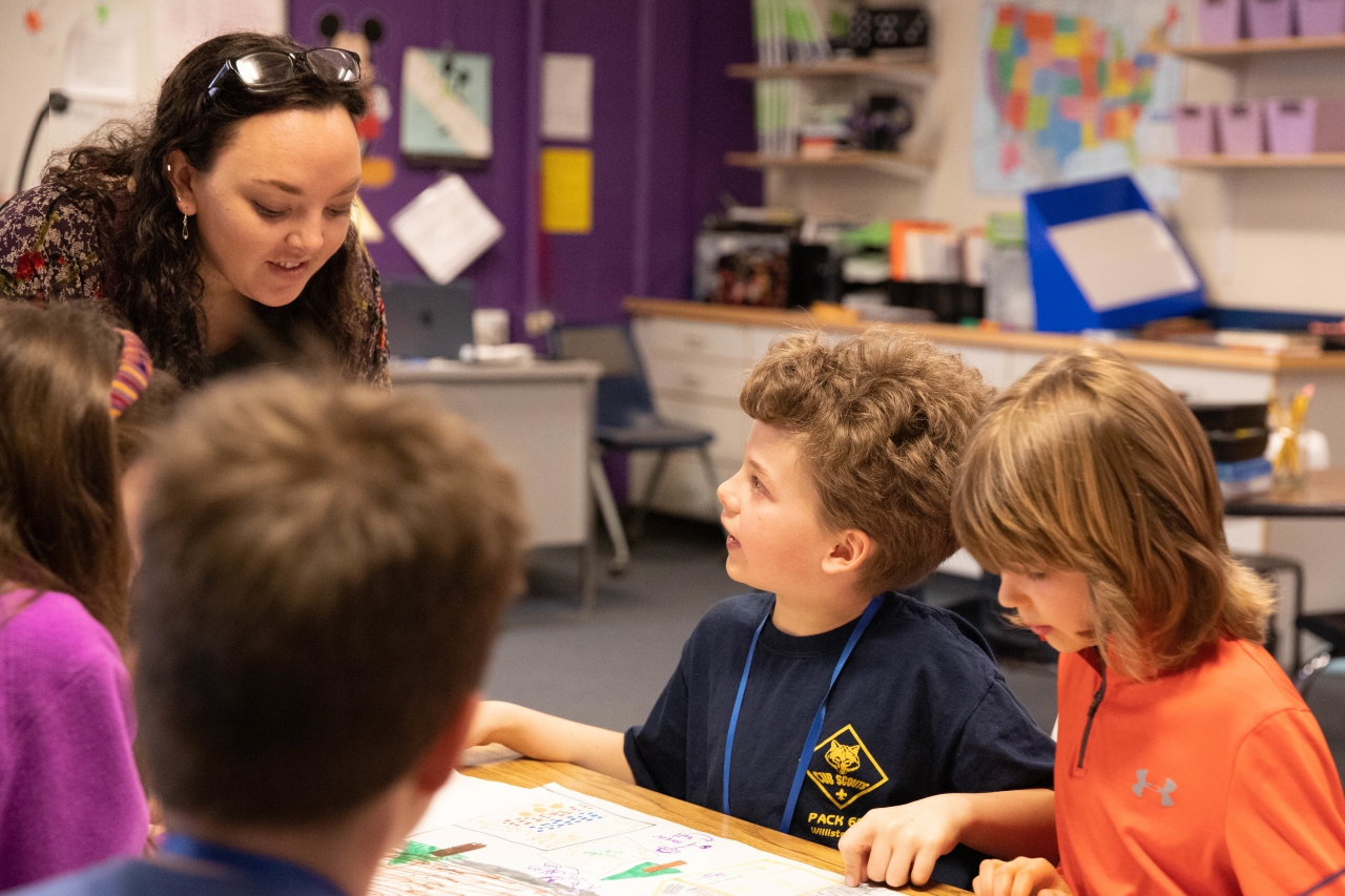 A teacher talks with of young students gathered around a table in a classroom. The table is covered with papers and drawings. 