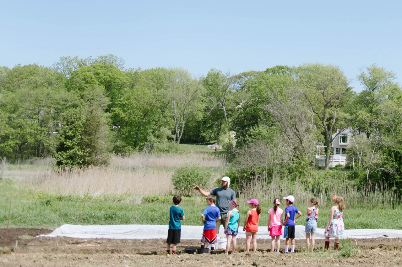 Students gather around a farmer in a field, trees and blue sky in the background.