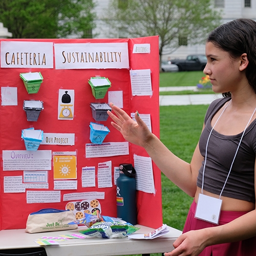 A young person gestures in front of a poster board display detailing cafeteria sustainability