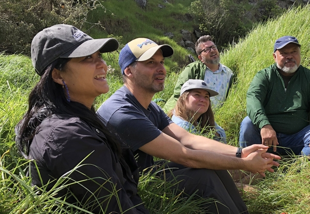 Five educators smile while listening intently, looking off camera. They are seated in tall grass with lush green mountains in the distance behind them.