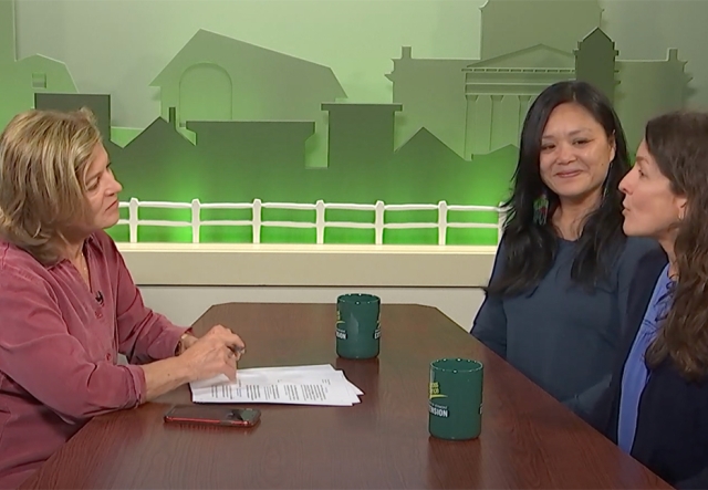 Three people sit around a desk in a television studio