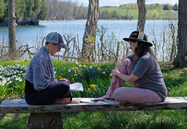 Two people sit on a bench, facing each other, deep in conversation, surrounded by green grass and spring flowers