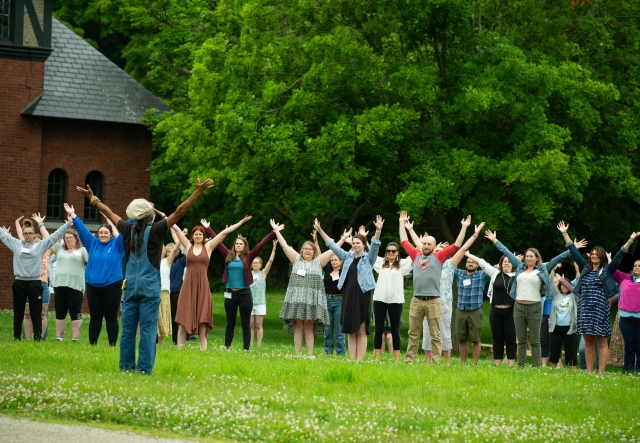 A large group of educators stand outdoors, raising their arms during a movement activity.