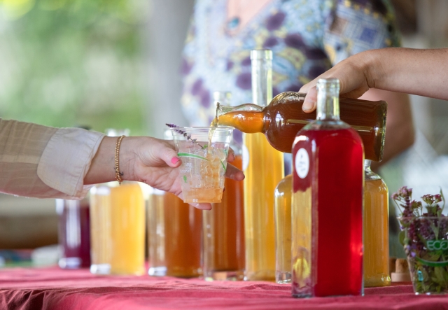 A person pours a beverage from a large bottle into a glass held by another person at a table with multiple bottles of colorful herbal syrups on a table.