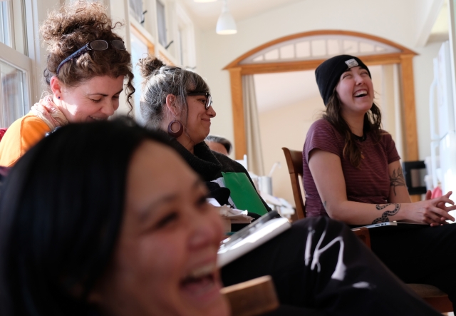 Four people sitting in chairs in a sunny living room laugh and smile