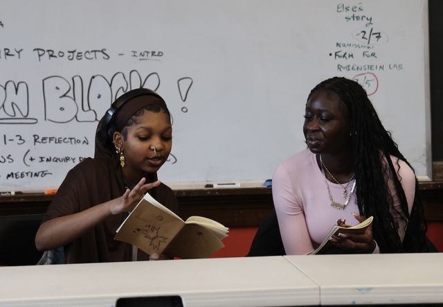 Two high school aged students discuss their journals while sitting in a classroom in front of a whiteboard
