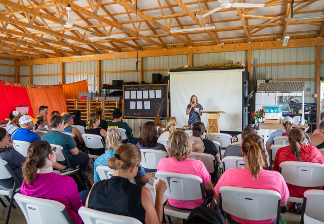 A person giving a presentation at a podium to an audience seated in a hall with educational materials displayed.