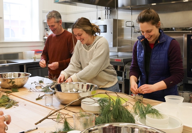 Three people smile while working with pine needles in an industrial kitchen