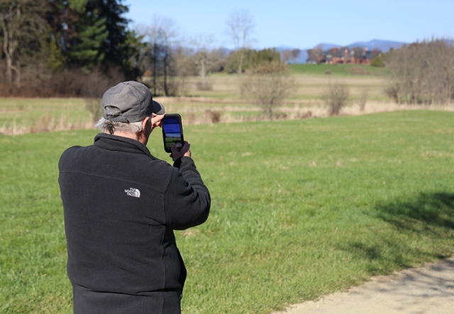 man facing away from camera taking a phone photo of field landscape