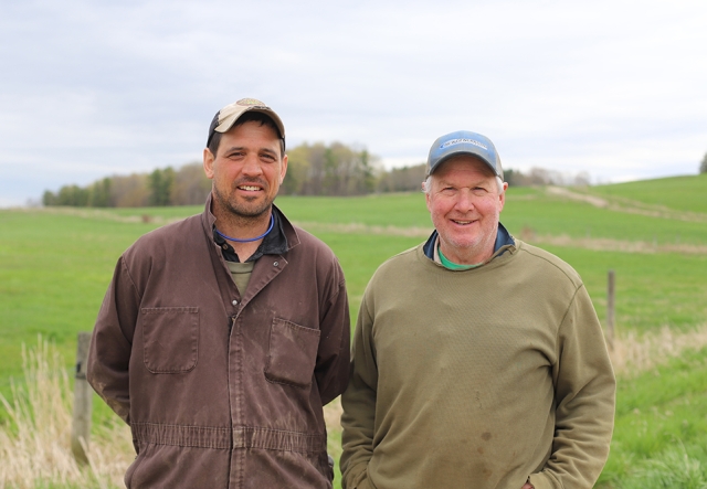 two men standing in front of a pasture. Man on the left is in a brown coverall and baseball cap. Man on right is gray-haired, also with a cap, and a ragged olive sweatshirt