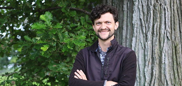 young man with dark curly hair and cropped beard standing with arms folded in front of big oak tree 
